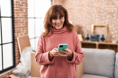 Middle age woman smiling confident using smartphone at new home
