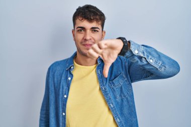 Young hispanic man standing over blue background looking unhappy and angry showing rejection and negative with thumbs down gesture. bad expression. 