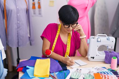 Middle age chinese woman tailor talking on smartphone writing on notebook at atelier