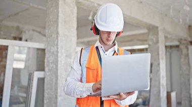 Kafkasyalı genç bir mimar, inşaat alanında dizüstü bilgisayar kullanarak rahat bir ifadeyle duruyor.