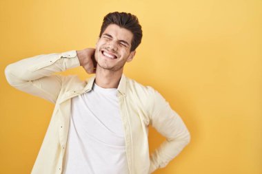 Young hispanic man standing over yellow background suffering of neck ache injury, touching neck with hand, muscular pain 