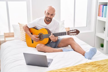 Genç kel bir adamın online klasik gitar dersi var. Yatak odasında oturuyor.