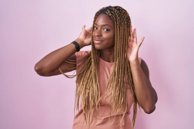 African american woman with braided hair standing over pink background trying to hear both hands on ear gesture, curious for gossip. hearing problem, deaf 