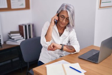 Orta yaşlı, kır saçlı, iş kadını. Akıllı telefondan konuşuyor. Ofise bakıyor.