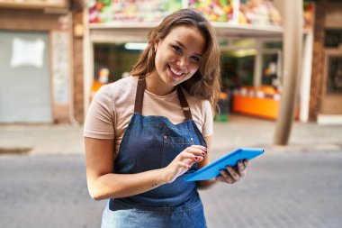Young beautiful hispanic woman waitress smiling confident using touchpad at fruit store