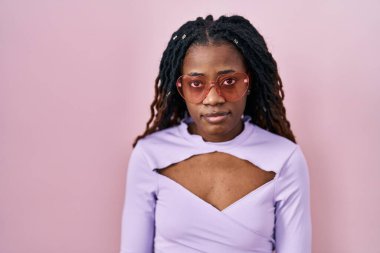 Pembe arka planda dikilen örülü saçlı Afrikalı kadın yüzünde ciddi bir ifadeyle rahatlamış. Basit ve doğal bir şekilde kameraya bakıyorsun.. 