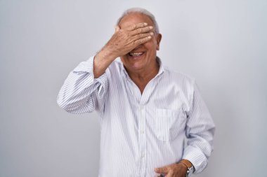 Senior man with grey hair standing over isolated background smiling and laughing with hand on face covering eyes for surprise. blind concept. 