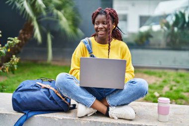African american woman student using laptop sitting on bench at campus park
