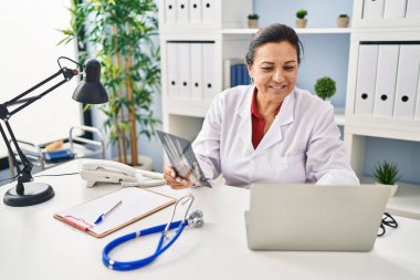 Middle age hispanic woman wearing doctor uniform holding xray at clinic