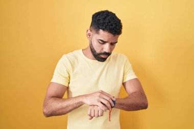 Sarı arka planda sakallı İspanyol bir adam kol saatinde saati kontrol ediyor, rahat ve kendinden emin. 