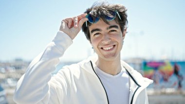 Genç İspanyol adam, deniz kenarında güneş gözlüğü takarak gülümsüyor.