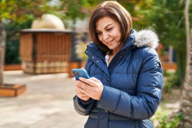 Parkta akıllı telefon kullanan orta yaşlı bir kadın gülümsüyor.