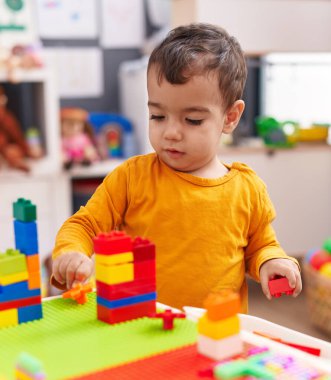 Adorable hispanic boy playing with construction blocks standing at kindergarten