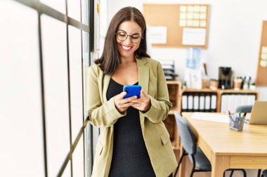 Genç, güzel İspanyol iş kadını ofisteki akıllı telefonu kullanarak gülümsüyor.