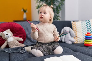 Sevimli sarışın kız anaokulu öğrencisi kanepede oturmuş defterine çizim yapıyor.