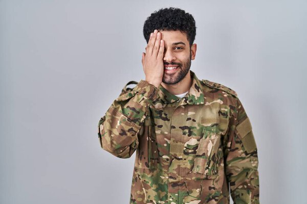 Arab man wearing camouflage army uniform covering one eye with hand, confident smile on face and surprise emotion. 