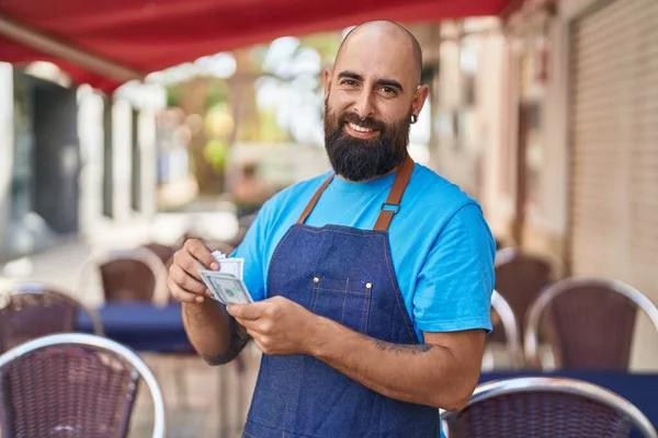 Young bald man waiter smiling confident counting dollars at coffee shop terrace