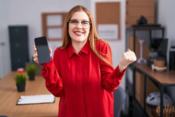 スマートフォンの画面を示すオフィスで働く赤毛の女性は誇りに思って叫んで 勝利と成功を祝う非常に腕を上げ興奮して — ストック写真
