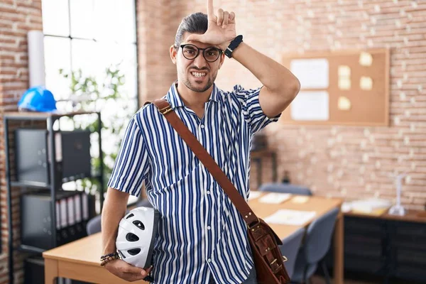 Hispanic man with long hair working at the office holding bike helmet making fun of people with fingers on forehead doing loser gesture mocking and insulting.