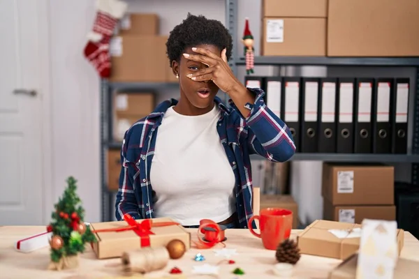 Stock image African american woman working at small business doing christmas decoration peeking in shock covering face and eyes with hand, looking through fingers with embarrassed expression. 