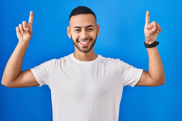 若いヒスパニック系の男は青の背景に立っている笑顔と驚きと指で指摘し 腕を上げ — ストック写真