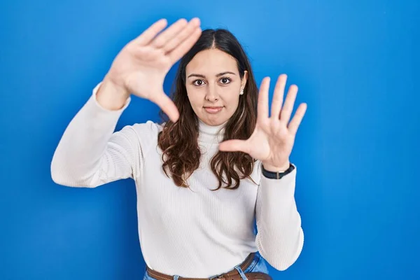 青い背景の上に立つ若いヒスパニック系の女性は 手のひらと指 カメラの視点を使用してフレームを行う — ストック写真