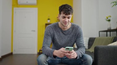 Young hispanic man using smartphone doing ok gesture at home