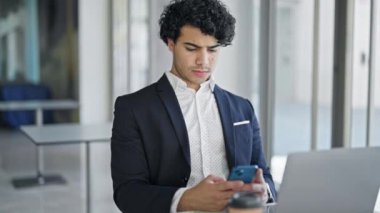 Genç Latin iş adamı laptopunu ve akıllı telefonunu kullanarak ofiste birinci sınıf jest yapıyor.