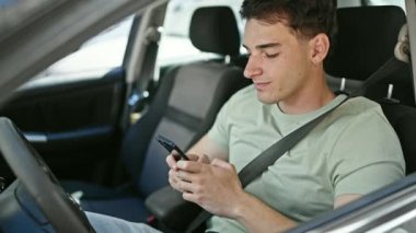Young hispanic man using smartphone sitting on car at street
