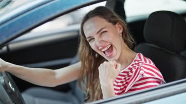 Genç sarışın kadın gülümsüyor. Kendine güvenen, kazanan yüz ifadesiyle arabada oturan.
