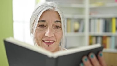 Kütüphanede gülümseyen, orta yaşlı, kır saçlı, kitap okuyan bir kadın öğrenci.