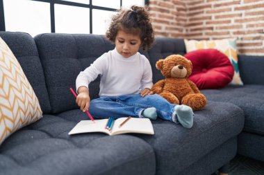 Adorable hispanic girl drawing on notebook sitting on sofa at home