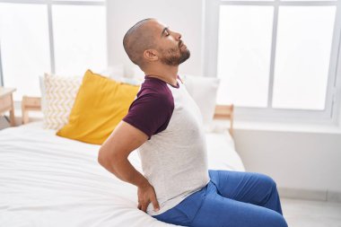 Young latin man suffering for back injury sitting on bed at bedroom