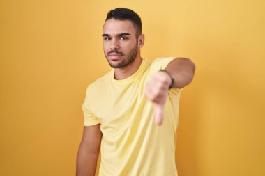 Young hispanic man standing over yellow background looking unhappy and angry showing rejection and negative with thumbs down gesture. bad expression. 