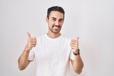 Beyaz arka plan başarı işaretinin üzerinde duran yakışıklı İspanyol adam elleriyle olumlu jest yapıyor, gülümsüyor ve mutlu. Neşeli ifade ve kazanan jest. 