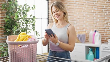 Young blonde woman using smartphone hanging clothes on clothesline at laundry room