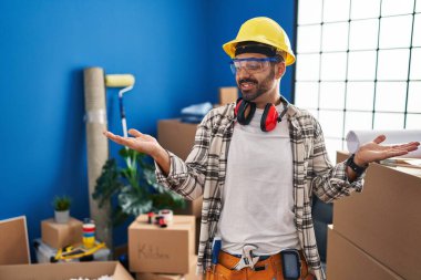 Evde çalışan sakallı genç İspanyol adam gülümseyerek iki elini de açık gösteriyor. Karşılaştırma ve reklam karşılaştırması yapıyor ve dengeyi sağlıyor. 