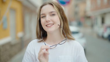 Young caucasian woman smiling confident holding glasses at street