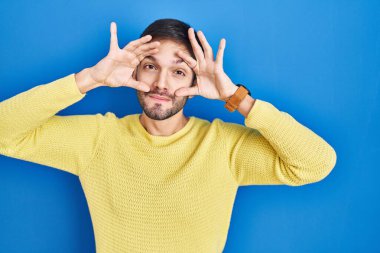 Hispanic man standing over blue background trying to open eyes with fingers, sleepy and tired for morning fatigue 