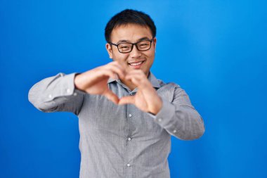 Mavi arka planda dikilen genç Çinli adam, aşkla gülümsüyor, elleriyle kalp sembolü yapıyor. romantik kavram. 