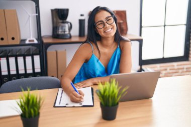 Young beautiful latin woman business worker using laptop writing on document at office