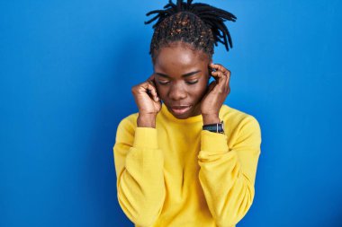 Beautiful black woman standing over blue background covering ears with fingers with annoyed expression for the noise of loud music. deaf concept. 