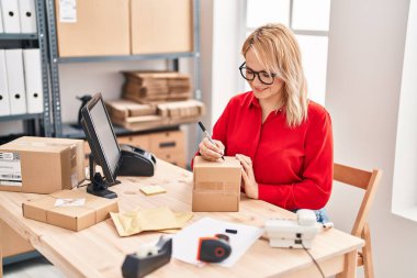 Genç sarışın kadın ecommerce iş işçisi ofiste paket üzerine yazıyor.