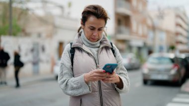 Orta yaşlı İspanyol kadın sokakta akıllı telefon kullanarak gülümsüyor.