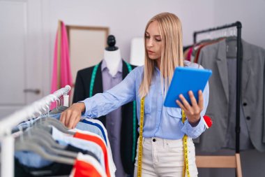 Young blonde woman tailor holding clothes on rack using touchpad at tailor shop