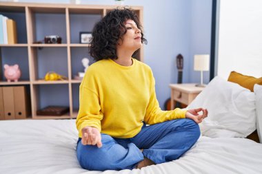 Young beautiful hispanic woman doing yoga exercise sitting on bed at bedroom clipart