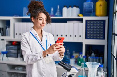 Young hispanic woman scientist smiling confident using smartphone at laboratory