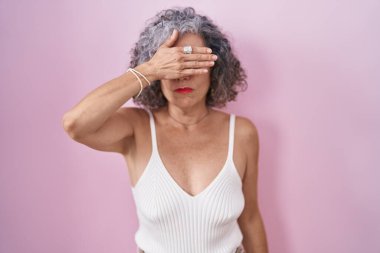 Middle age woman with grey hair standing over pink background covering eyes with hand, looking serious and sad. sightless, hiding and rejection concept 