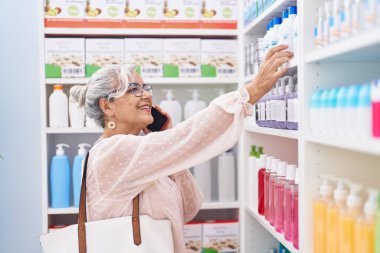 Orta yaşlı, gri saçlı, gülümseyen bir müşteri eczanede akıllı telefondan konuşuyor.