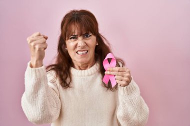 Middle age hispanic woman holding pink cancer ribbon annoyed and frustrated shouting with anger, yelling crazy with anger and hand raised 